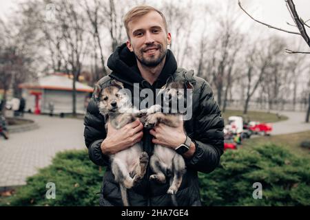 Young man holds and carries two small wolf-like puppies in his arms. Stock Photo