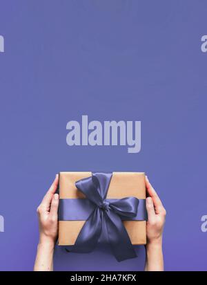 Female hands hold gift box on very peri violet color background with copy space. Caucasian hands holding gift box in craft wrapping paper with fashion color 2022 very peri satin ribbon. Vertical. Stock Photo