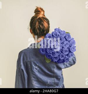 Back view of young woman in denim jacket holding bunch of violet roses on shoulder. Girl with bun updo in jeans holding flowers in very peri color 2022. Copy space. Demonstrating color of 2022 year Stock Photo