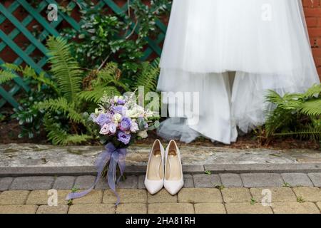 Wedding details: classic bride shoes and bouquet of roses standing on the background pf dress and greenary. Very Peri - trendy color of the year 2022 in weddings flowers.  Stock Photo