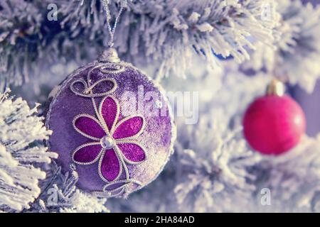Snowy branch of christmas tree with violet gray fabric christmas ball with flower and glitter decorations close up and blurred red ball on background. Stock Photo