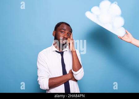 Professional businessman thinking under white paper thought bubble concept standing with arms crossed and finger pointing at head. African american entrepreneur brainstorming next to idea cloud. Stock Photo