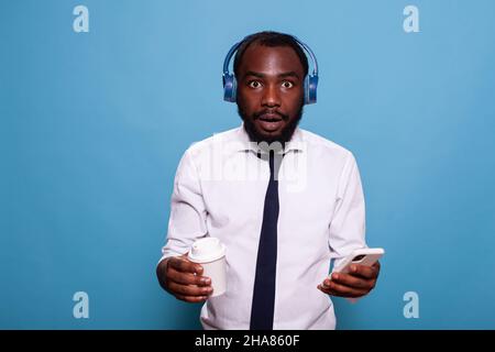 Shocked man listening to music on wireless headphones while holding smartphone and cup of coffee to go. Businessman looking surprised streaming audio content on over the ear headset. Stock Photo