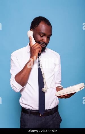 Calm businessman holding reciever to ear standing up talking on landline cord telephone listening to person speaking at the other end of the line. Employee in white shirt using oldschool phone. Stock Photo