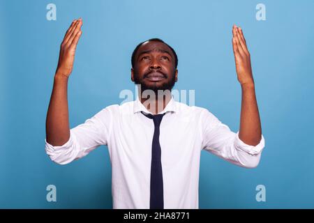 Worried businessman praying looking up with hands raised toward the sky acting humble. Emotional employee in white shirt with black tie asking for forgiveness. Stock Photo