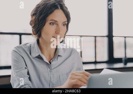 Young female office worker sitting at workplace and working online on laptop, Stock Photo