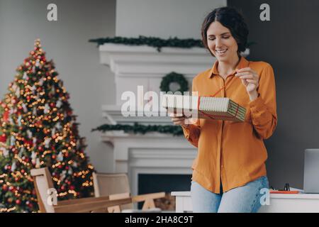 Happy european woman opening gift while standing in gorgeous Christmas decorated living room Stock Photo