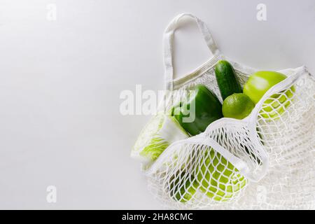 Green fruits and vegetables in white string bag. Green apples, lime, cucumber, bell peppers and cabbage in a reusable string cotton bag on a white bac Stock Photo