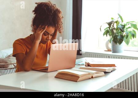 Tired flustrated African descent young girl sitting at desk in front of laptop while irritably looking at computer screen with hands holding her head Stock Photo