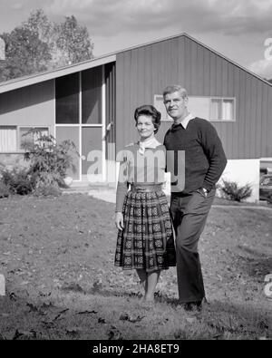 1950s 1960s MAN AND WOMAN ARM IN ARM STANDING IN FRONT OF THEIR MID-CENTURY MODERN HOME  - s12423 HAR001 HARS PROUD MARRIED SPOUSE HUSBANDS MODERN HOME LIFE COPY SPACE FRIENDSHIP FULL-LENGTH LADIES PERSONS RESIDENTIAL MALES BUILDINGS MIDDLE-AGED B&W PARTNER MIDDLE-AGED MAN EYE CONTACT GOALS STRUCTURE HAPPINESS MIDDLE-AGED WOMAN PROPERTY AND PRIDE SPLIT LEVEL HOMES REAL ESTATE RESIDENCE STYLISH HOMEOWNERS MID-ADULT MID-ADULT MAN MID-ADULT WOMAN TOGETHERNESS WIVES BLACK AND WHITE CASUAL CAUCASIAN ETHNICITY HAR001 MID-CENTURY OLD FASHIONED Stock Photo