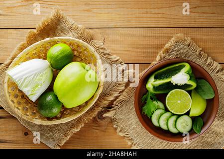 Various green fruits and vegetables on a wooden table. Chopped green apple, bell pepper, cucumber, cabbage and lime in a clay bowl. Top view Stock Photo