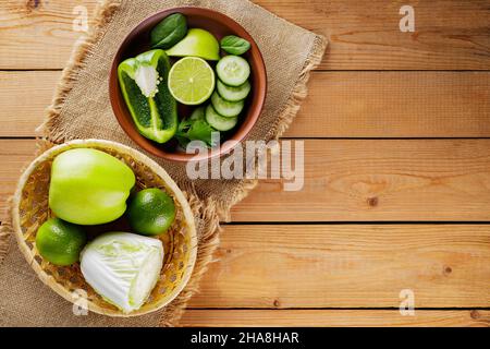 Various green fruits and vegetables on a wooden table. Chopped green apple, bell pepper, cucumber, cabbage and lime in a clay bowl. Top view Stock Photo