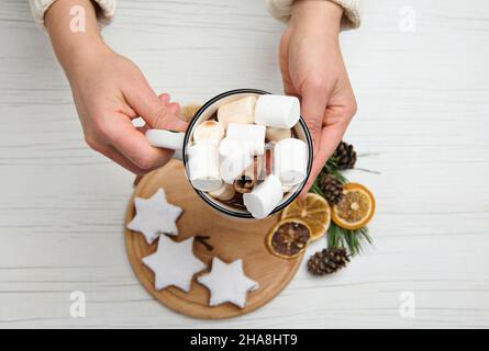 Hands holding a cup of hot chocolate drink with marshmallows over a wooden board with star-shaped gingerbread cookies sprinkled with powdered sugar an Stock Photo