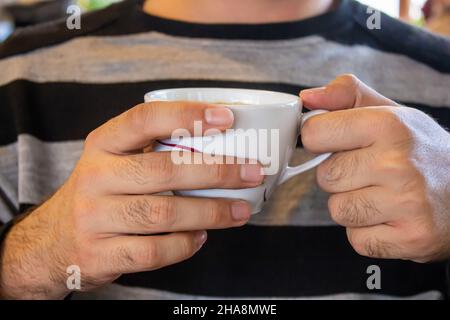 Closeup photo of young man holding cup of coffee while sitting in a modern cafe. Stock Photo