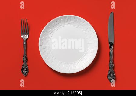 Empty white dinner plate, fork and table knife against red background. Blank mockup template for unhealthy eating, fast foods and bad eating habits. Stock Photo