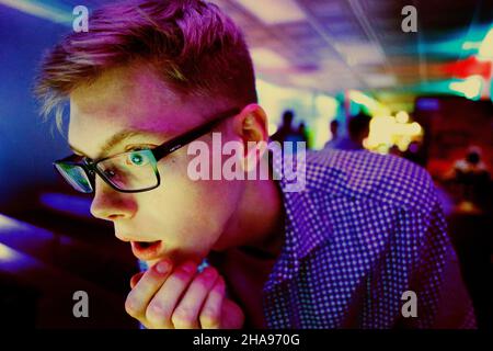 teen male playing video game in arcade. young adult in awe. neon lights flash in boy's face. young man in glasses with shocked expression. Stock Photo