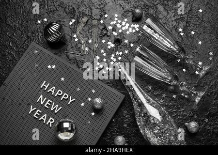 Board with text HAPPY NEW YEAR, glasses, opened champagne bottle and Christmas decor on black background Stock Photo