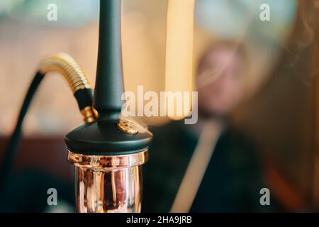 Hookah on table in lounge bar. Smoking person in blurry background. Close-up of black pipe and golden shisha flask. Leisure in oriental cafe. Stock Photo