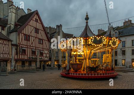 An illuminated Christmas children's carousel turns in front of a gable-fronted half-timbered house on the square in Dijon, Burgundy, France. Passers-by cross the cobblestones in the twilight, with strings of lights stretched between the roofs. Stock Photo