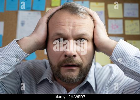 Surprised Middle-aged Holding his head on hands and looking at the camera. Task Sticky notes on background. Humorous portrait Stock Photo