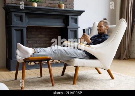Happy carefree millennial cellphone user relaxing in armchair at home Stock Photo