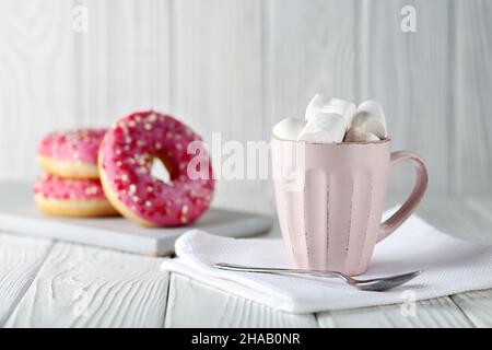 Donuts with pink glaze and colored caramel powder and cocoa with marshmallows on a white wooden background. Stock Photo