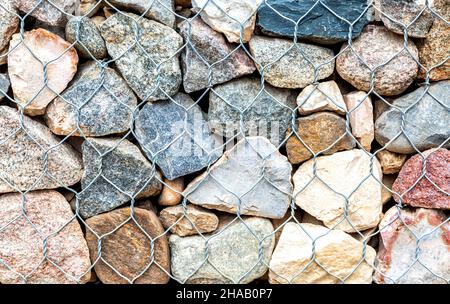 Stone wall with metal grid and rough stones as background. Rock texture in metal grid. Gabion net with stones Stock Photo