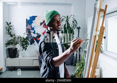 Inspired happy black man painting on an easel inside of his apartment. Lots of potted plants and an abstract painting on the wall. Stock Photo