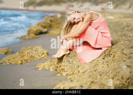 Mature woman sitting on some rocks on the shore of a tropical beach, wearing a nice orange dress. Stock Photo