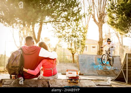 Multiracial couple in love sitting at skate park with music watching friends on bmx freestyle exhibition - Urban relationship concept Stock Photo