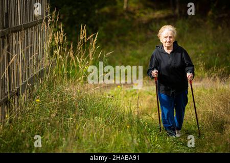 An elderly woman walks with sticks, practicing Nordic walking in the countryside. Stock Photo