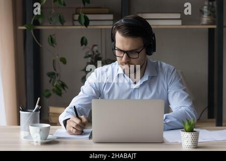 Man in headphones sit at desk holds pen noting information Stock Photo