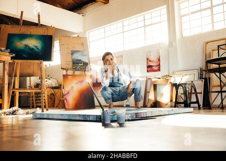 Happy young painter squatting on the floor while working in her art studio. Female artist smiling at the camera and holding a paintbrush. Creative you Stock Photo