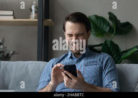 Man sits on sofa holds smartphone laughs on funny video Stock Photo