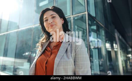Carefree businesswoman smiling at the camera while standing in front of a high rise office building in the city. Successful female entrepreneur standi Stock Photo