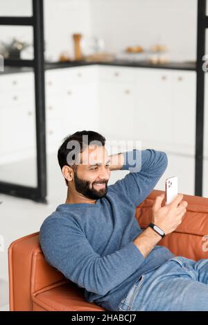 Vertical shot of Indian man resting on the comfortable sofa at home, using mobile phone for spending leisure, happy hispanic latin guy scrolling feeds, enjoying online chatting Stock Photo