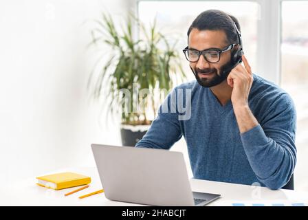Smiling Indian office worker in glasses and headset looking at laptop screen and talking online, positive ethnic man holding video call, working in customer service department, explaining something Stock Photo