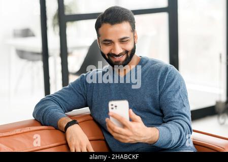 Headshot of Indian man resting on the comfortable sofa at home, using mobile phone for spending leisure, happy hispanic latin guy scrolling feeds, enjoying online chatting Stock Photo