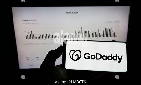 Person holding mobile phone with logo of American web hosting company GoDaddy Inc. on screen in front of web page. Focus on phone display. Stock Photo