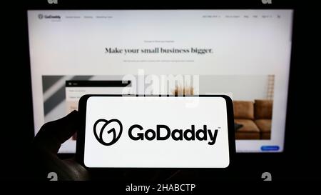 Person holding smartphone with logo of US web hosting company GoDaddy Inc. on screen in front of website. Focus on phone display. Stock Photo