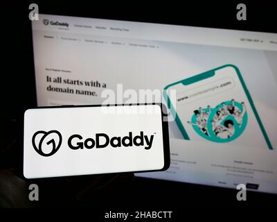 Person holding cellphone with logo of US web hosting company GoDaddy Inc. on screen in front of business webpage. Focus on phone display. Stock Photo