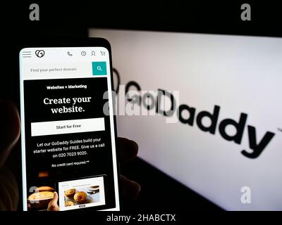 Person holding smartphone with website of American web hosting company GoDaddy Inc. on screen in front of logo. Focus on center of phone display. Stock Photo