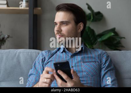 Man sits on sofa holds smartphone looking into distance Stock Photo