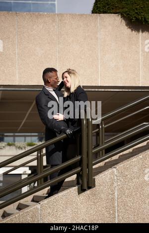 Happy black man and blond woman embracing each other and smiling while standing on stairway on modern city street in daytime Stock Photo