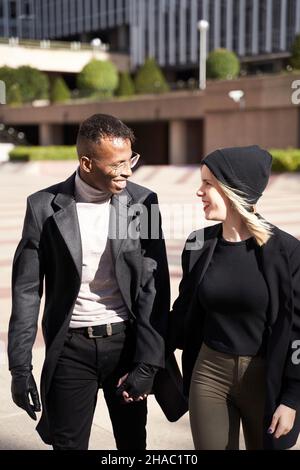 Happy multiethnic man and woman in casual clothes holding hands and looking at each other while strolling on city street during romantic date, on sunny day Stock Photo