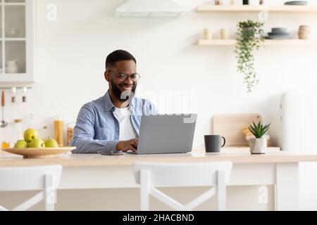 Smiling millennial african american bearded guy in glasses working on laptop in minimalist kitchen interior Stock Photo