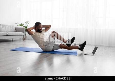 Happy young african american guy doing exercises for legs and abs on mat on floor in room interior Stock Photo