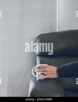 A female's hand holding a white cup on the armrest on an armchair Stock Photo