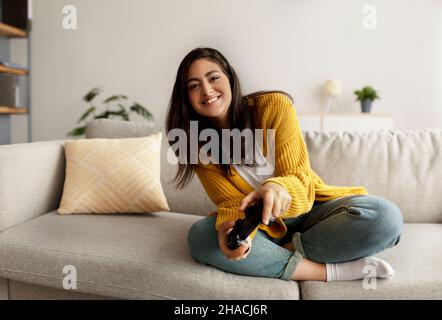 Fun stay home pastimes. Joyful middle eastern woman using joystick to play online video games, sitting on sofa Stock Photo
