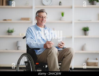Cheerful senior man in wheelchair using tablet computer, browsing internet at home Stock Photo